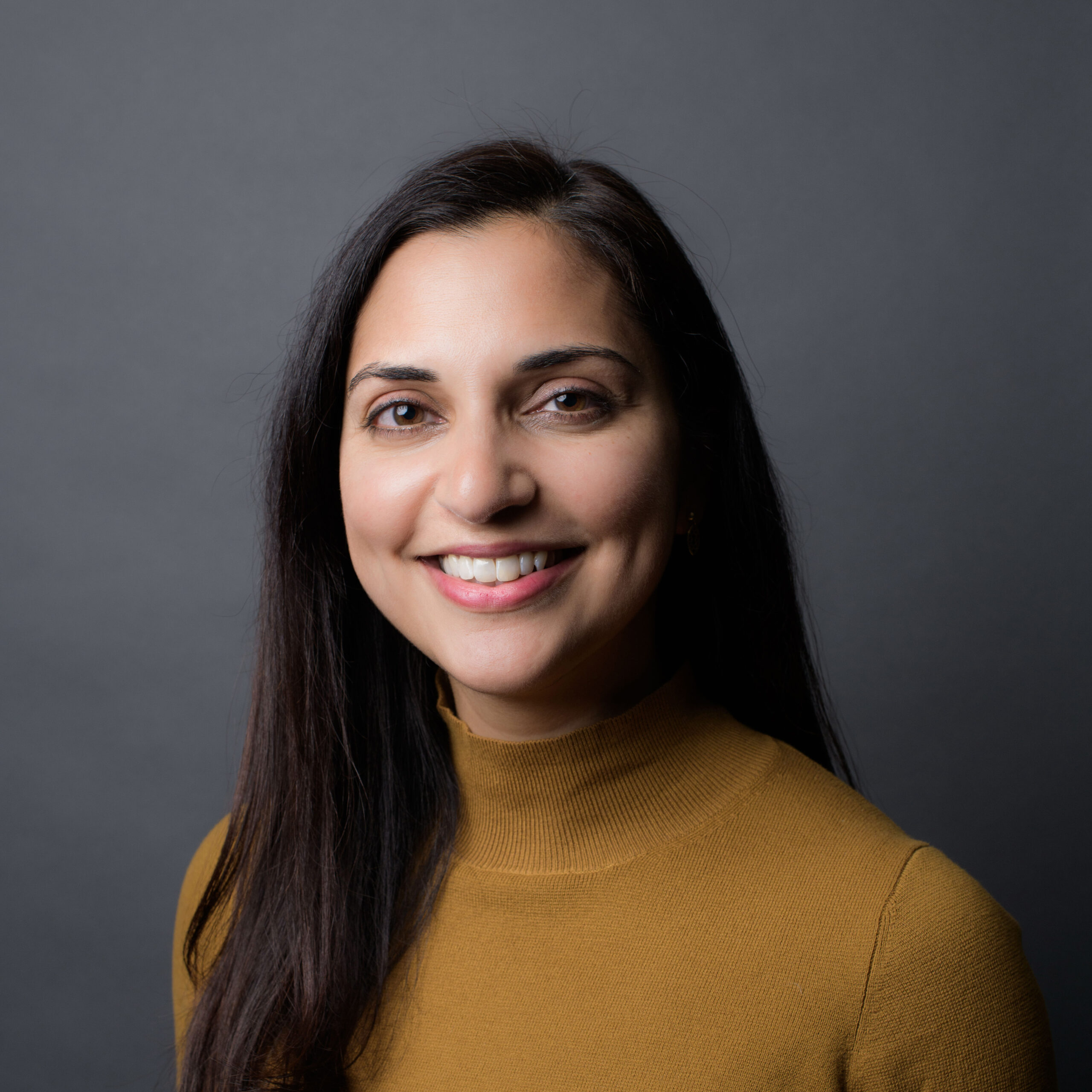 Medha Makhlouf. A woman with long black hair smiles at the camera. She is wearing a gold turtleneck sweater.